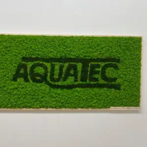Machový obraz so svetlozeleným lišajníkom a tmavozeleným logom AQUATEC v svetlom drevenom ráme na bielej stene.